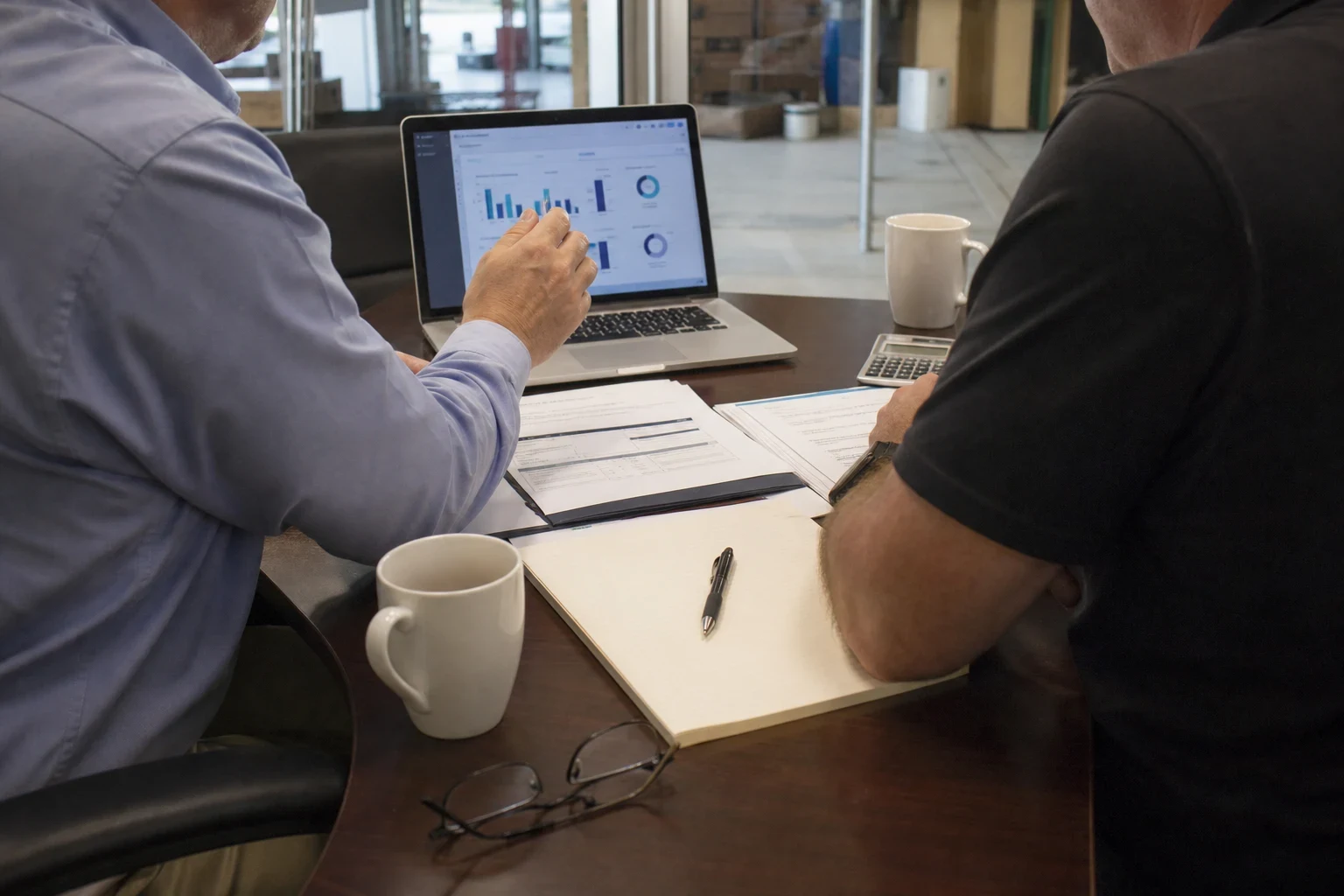 Two men seated at a round wooden table reviewing charts on a laptop, with paperwork, a notepad, coffee mugs, a calculator and glasses, near a glass storefront.
